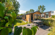 Acogedora cabaña con terraza y muebles de jardín, rodeada de otras casas y vegetación en un día soleado.