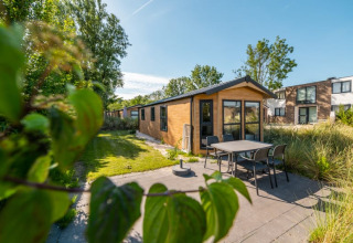Acogedora cabaña con terraza y muebles de jardín, rodeada de otras casas y vegetación en un día soleado.