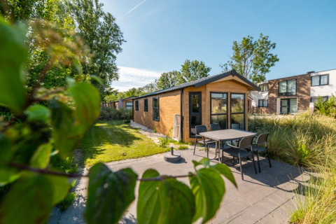 Acogedora cabaña con terraza y muebles de jardín, rodeada de otras casas y vegetación en un día soleado.