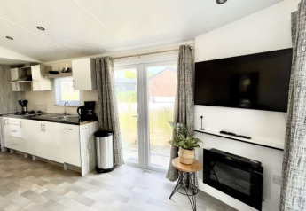 Intérieur de lodge moderne avec cuisine ouverte, TV murale, cheminée, grandes fenêtres et déco soignée.