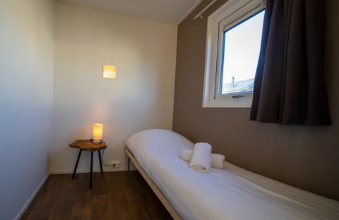 A cozy lodge room with a single bed, rolled towels, window with curtains, and a lamp on a small table.