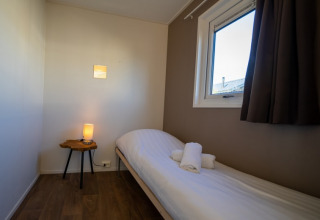 Petite chambre confortable d’un lodge avec lit simple, fenêtre, rideaux, et lampe posée sur une table.
