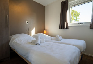 Bright lodge room with two single beds, clean white linens, towels, a window, and brown curtains.