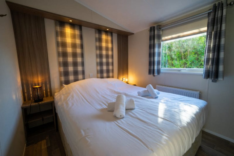 Cozy lodge bedroom with double bed, plaid curtains, warm lighting, and a window facing lush greenery.