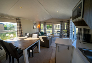Intérieur de lodge moderne avec table à manger, canapé, coin cuisine et grandes fenêtres sur la nature.