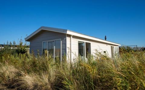 Petit lodge clair entouré de hautes herbes et de végétation sous un ciel bleu limpide, journée ensoleillée.