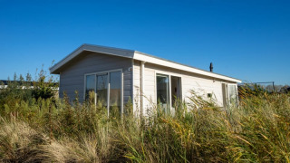 Petit lodge clair entouré de hautes herbes et de végétation sous un ciel bleu limpide, journée ensoleillée.