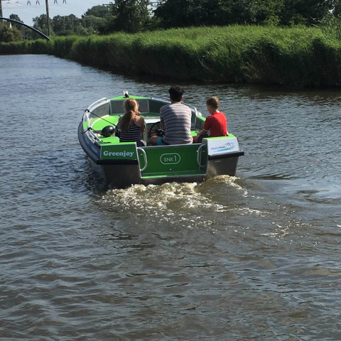 Tres personas navegan en una pequeña lancha verde llamada Greenjoy por un canal tranquilo cerca de Loënga, Friesland.