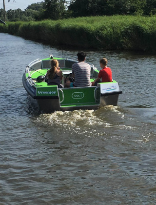 Tres personas navegan en una pequeña lancha verde llamada Greenjoy por un canal tranquilo cerca de Loënga, Friesland.