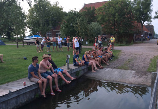 Folk sidder og slapper af ved vandet på Camping Pasveer i Friesland, Holland med grønne omgivelser.