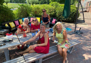Cinque donne sorridono sedute a un tavolo da picnic al Camping Pasveer in Frisia, Paesi Bassi, in una giornata soleggiata.