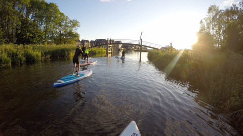 Folk står på paddleboards på en rolig kanal ved Camping Pasveer i Friesland, Holland, med solnedgang.