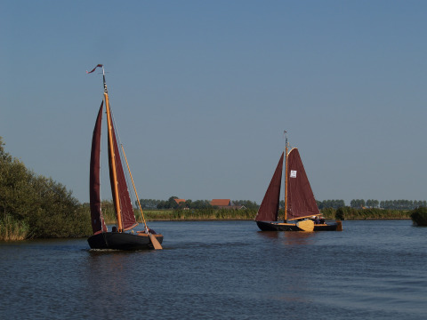Dos veleros de velas rojas navegan por un río tranquilo cerca de Loënga, Frisia, Países Bajos bajo cielo azul.