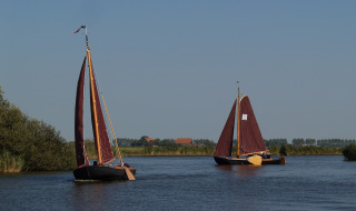 Dos veleros de velas rojas navegan por un río tranquilo cerca de Loënga, Frisia, Países Bajos bajo cielo azul.