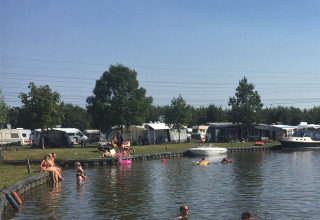 Feriecamping ved sø i Friesland, Nederlandene, med svømmende mennesker, telte og både under blå himmel.