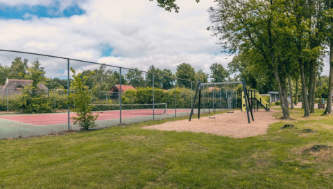Legeplads og tennisbane ved en feriepark med glamping, omgivet af træer og grønne områder.