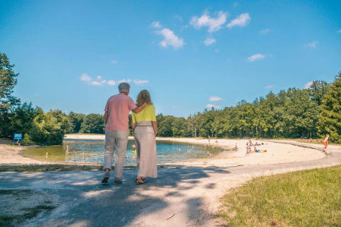 Older couple walking arm in arm by a lake in a holiday park, with families relaxing on a sandy beach.