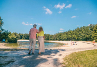 Older couple walking arm in arm by a lake in a holiday park, with families relaxing on a sandy beach.