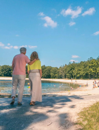 Pareja mayor paseando abrazada junto a un lago en un parque vacacional con personas disfrutando la playa.