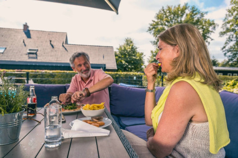 An older couple enjoys an outdoor meal at a table in a glamping holiday park with a green background.