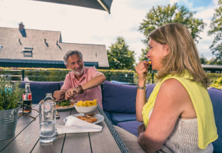 Een ouder koppel geniet van een maaltijd op het terras in een glamping vakantiepark met groene achtergrond.