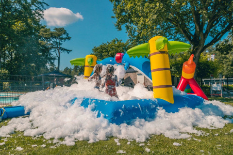 Enfants jouant dans la mousse sur une structure gonflable avec palmiers dans un parc de vacances glamping.