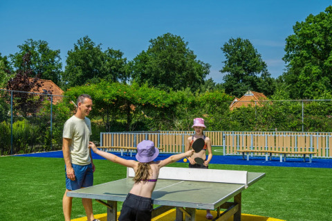 Dos niños y un adulto juegan tenis de mesa al aire libre en un parque vacacional con alojamiento glamping.