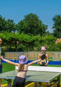 Dos niños y un adulto juegan tenis de mesa al aire libre en un parque vacacional con alojamiento glamping.