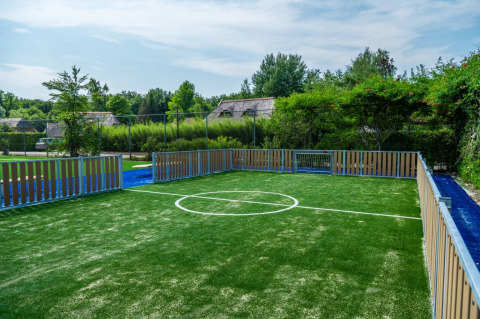 Moderne minifodboldbane med hegn og kunstgræs i en feriepark omgivet af grønne træer og hytter.