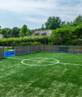 Pequeña cancha de fútbol con césped artificial y vallas en un parque vacacional rodeado de naturaleza.