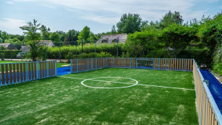 Pequeña cancha de fútbol con césped artificial y vallas en un parque vacacional rodeado de naturaleza.