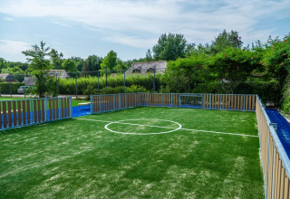 Modern mini football pitch with artificial turf and fencing at a holiday park surrounded by greenery.