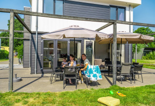 Gezin ontspant onder parasols op het terras van een modern glampingverblijf in een vakantiepark.