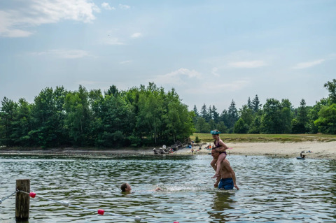 Menschen schwimmen in einem See an einem sandigen Ufer eines Ferienparks mit Glamping-Angeboten.