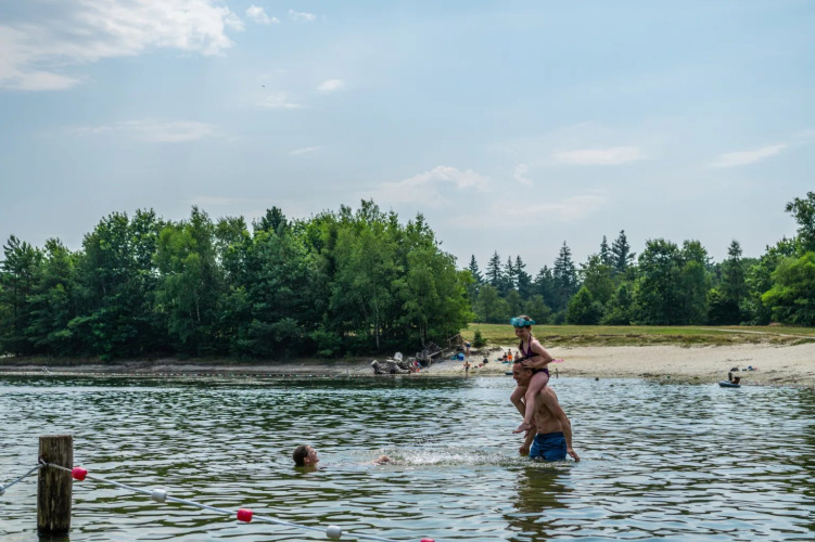 People swimming and playing in a lake near a sandy beach at a holiday park offering glamping options.