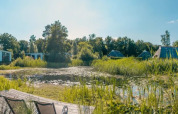 Panoramabillede af glampinghytter ved en sø omgivet af grøn natur i en feriepark under blå himmel.