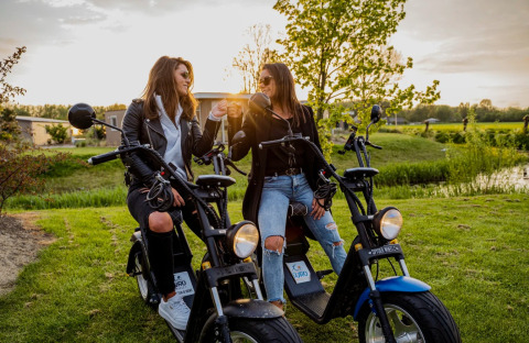 Twee vrouwen zitten op elektrische scooters in een vakantiepark tijdens zonsondergang, genietend van glamping.