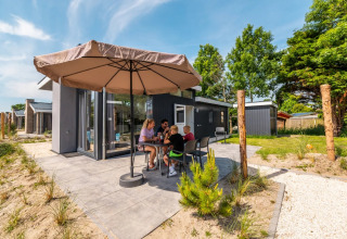 Familie sitzt unter Sonnenschirm und genießt eine Mahlzeit vor einer modernen Glamping-Unterkunft.