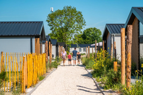 Family walking on a gravel path between modern glamping cabins surrounded by greenery in a holiday park.