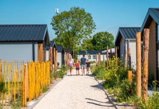 Familie går på grussti mellem moderne glampinghytter og grønne planter i en feriepark på en solrig dag.