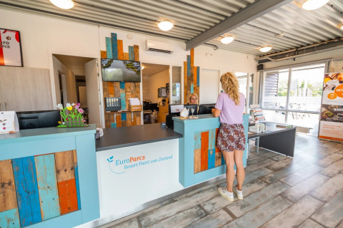 Receptie van een vakantiepark met een vrouw aan de balie, kleurrijke details en zomerse sfeer.