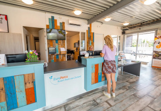 Receptie van een vakantiepark met een vrouw aan de balie, kleurrijke details en zomerse sfeer.