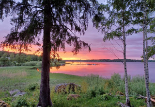 Sunset view at Camping Falkudden, Dalarna, Sweden with a calm lake, grassy shore, and silhouetted trees.