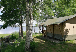 Safari tent by the lakeside, nestled among green trees on a sunny day, perfect for a nature getaway.