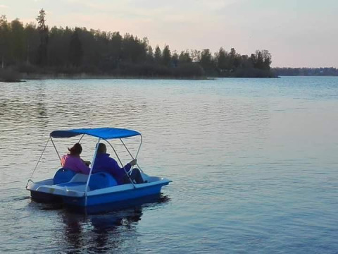 Two people ride a pedal boat on a calm lake at Camping Falkudden, surrounded by trees in Dalarna, Sweden.