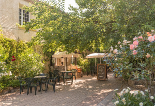 Udendørs caféområde med borde, stole og parasoller omgivet af træer og blomster i feriepark i Occitanie.