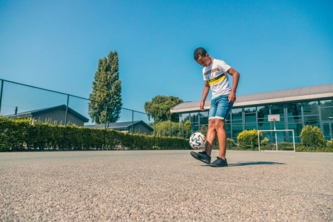 Jongen speelt met een voetbal op een buitenplein bij vakantiepark met glampingaccommodaties en helder weer.