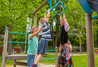 Kinderen spelen op een klimrek in een vakantiepark, begeleid door volwassenen, omringd door groen.