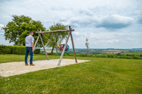 Man duwt kinderen op een schommel in een open weide bij een glamping vakantiepark met panoramisch uitzicht.