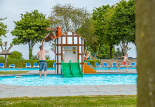 Buitenzwembad met speeltoestellen in een vakantiepark, enkele kinderen en volwassenen genieten van het water.